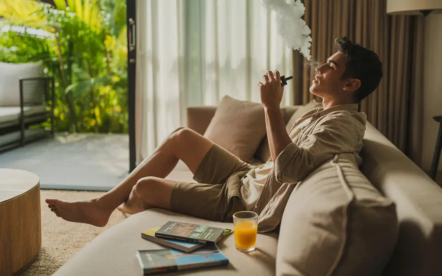 A tourist sitting on a sofa and vaping cannabis in a private, tropical accommodation in Thailand.
